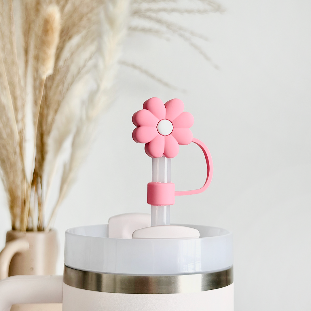 A close-up photo showcasing Coconut Lane's Pink & White Daisy Straw Cover attached to a straw, nestled inside a white cup with a silver rim. In the background, there is a blurred arrangement of dried pampas grass in a beige vase, creating a minimalistic and bright scene.
