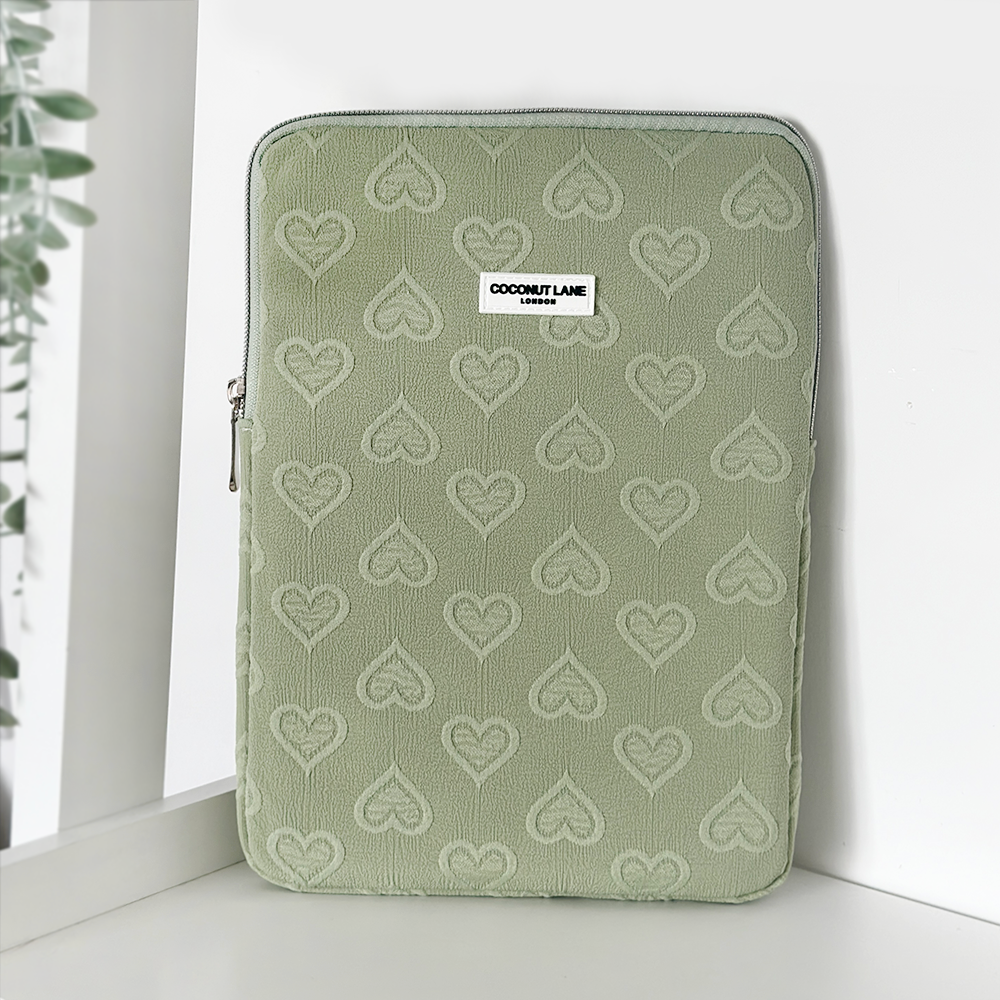 A Sage Heart iPad Sleeve with a zip closure from Coconut Lane, featuring an embossed heart pattern. The sleeve is standing upright against a white background with a small part of a plant visible on the left side. A white label with "Coconut Lane" is sewn on the front. Perfectly sized for 11" iPads, this versatile case doubles as an iPad Sleeve.