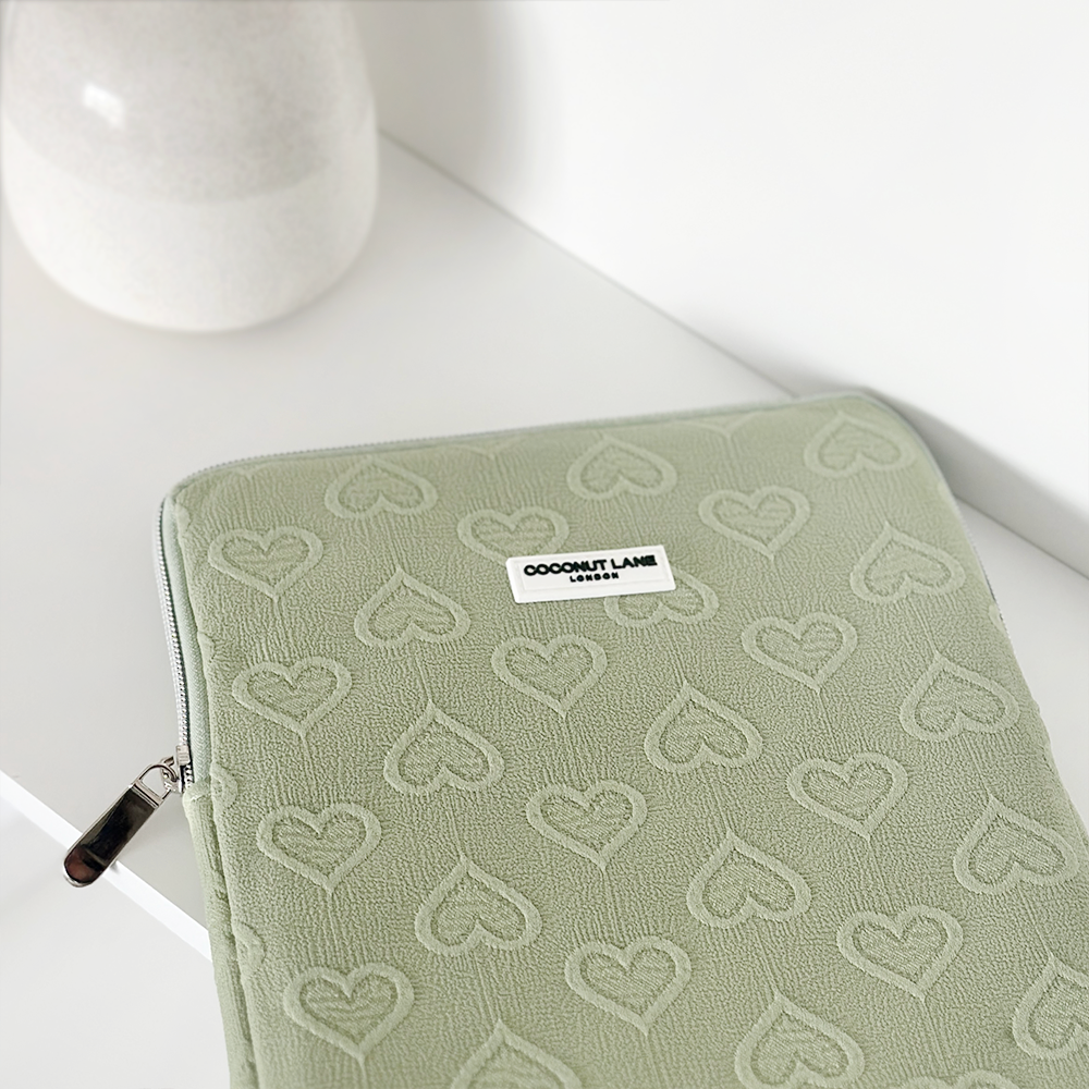 A Sage Heart iPad Sleeve from Coconut Lane, designed for 11" iPads, rests on a white surface. The sage green zippered pouch features a light heart pattern and a small label that reads "COCONUT LANE LONDON." In the background, a white ceramic vase can be partially seen on the left side.