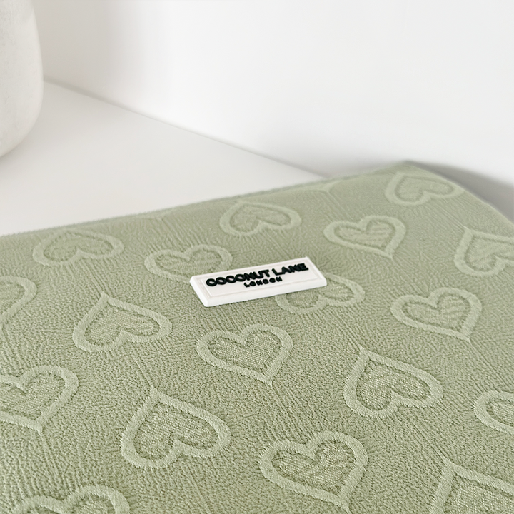 The Sage Heart iPad Sleeve from Coconut Lane features a textured green fabric adorned with an embossed heart pattern, perfectly fitting 11" iPads. A white label with black text reading "COCONUT LANE, LONDON" is sewn onto the fabric. The background is a clean, white surface.