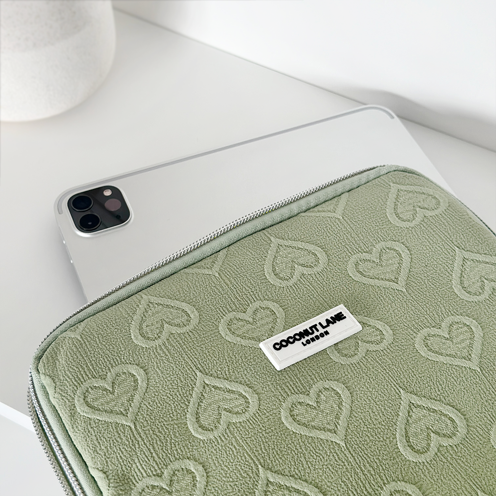 A white tablet, partially inside a Sage Heart iPad Sleeve designed for 11" iPads, is placed on a white surface. The light green pouch is adorned with embossed heart patterns and has a label that reads "Coconut Lane." A white cylindrical object is slightly visible in the background.