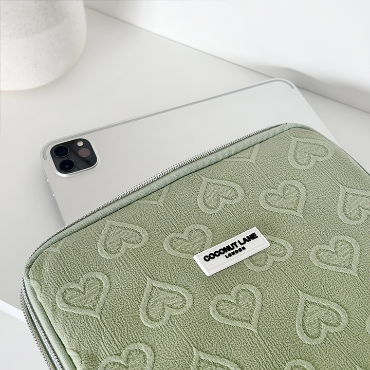 A white tablet, partially inside a Sage Heart iPad Sleeve designed for 11" iPads, is placed on a white surface. The light green pouch is adorned with embossed heart patterns and has a label that reads "Coconut Lane." A white cylindrical object is slightly visible in the background.