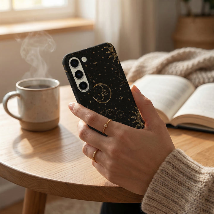 Hand holding a navy blue celestial-patterned Samsung phone case near an open book and steaming mug on a wooden table.