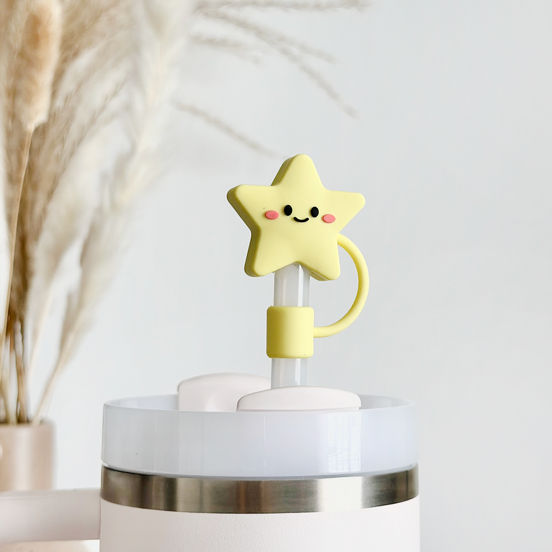 The Star Straw Cover by Coconut Lane is an adorable yellow, star-shaped silicone decoration with a cute smile and rosy cheeks. It is attached to a white and silver cylindrical object and photographed in front of a blurred background adorned with dried pampas grass.