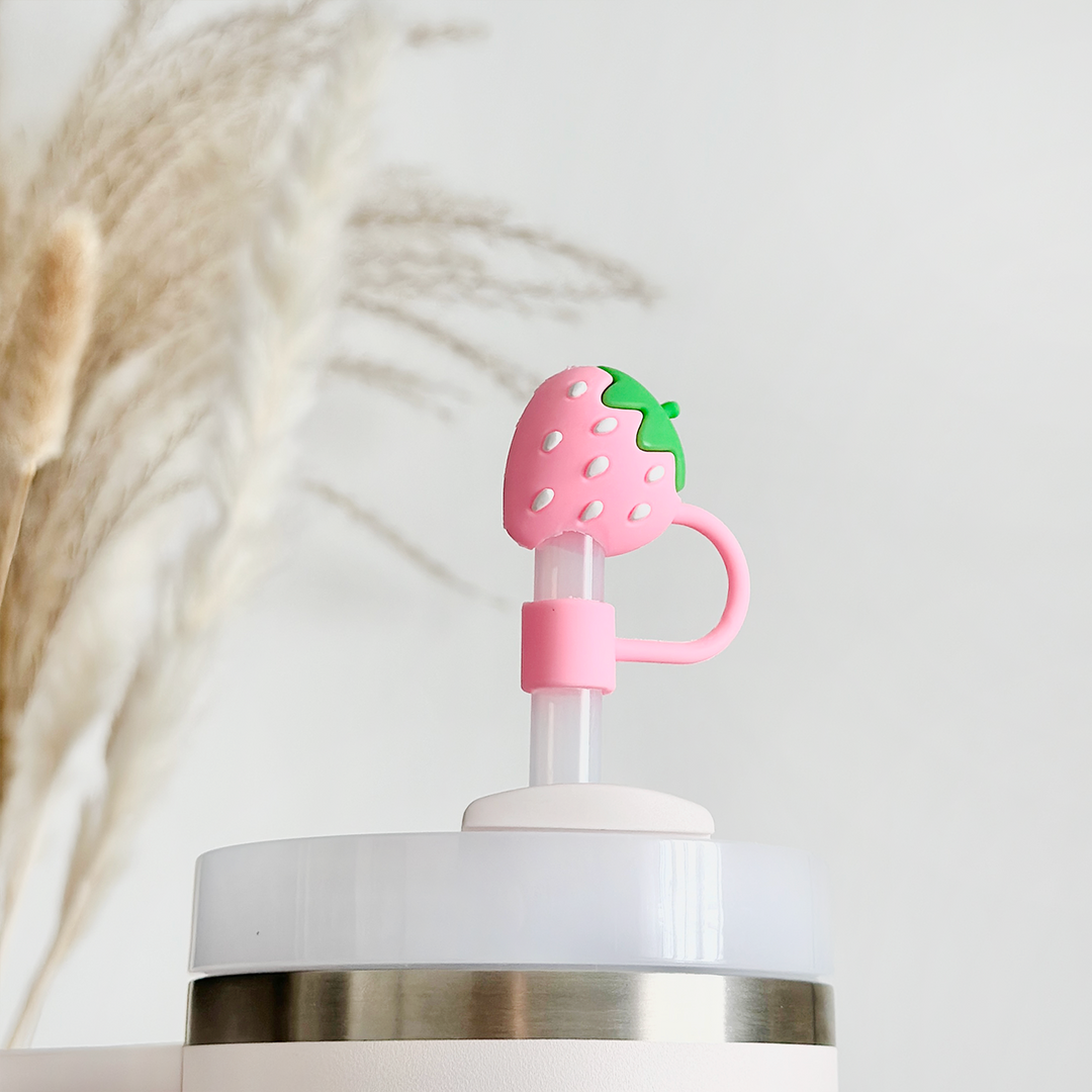 A close-up of a cup with a straw featuring Coconut Lane's Strawberry Straw Cover, a decorative pink strawberry-shaped silicone topper. The background is blurred, showing dry pampas grass against a light-colored wall.