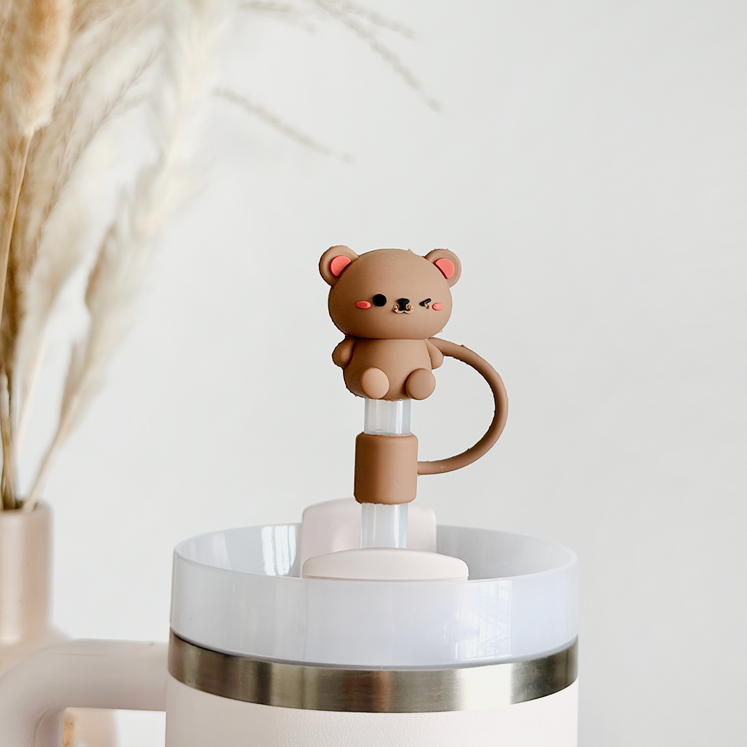 A Teddy Bear Straw Cover from Coconut Lane, shaped like a cute brown bear with a long tail and pink blush on its cheeks, sits atop a neutral-colored silicone straw in a white cup, surrounded by a blurred background of soft beige pampas grass.