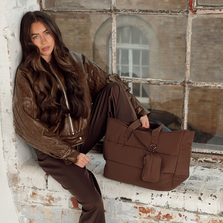 Woman with long brunette hair sat on a industrial brick window sill setting. Next to her is the Coconut Lane Main Carry choc brown puffer bag. Attached on the external d-ring is the matching mini keyring bag. 