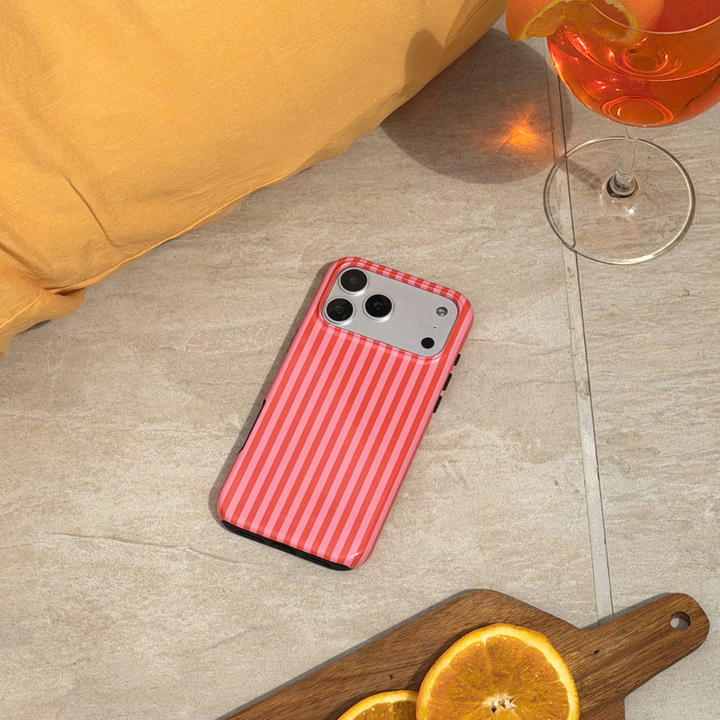 Phone with a pink and orange striped case on a tiled floor next to a glass of orange aperol spritz and a cutting board with oranges.