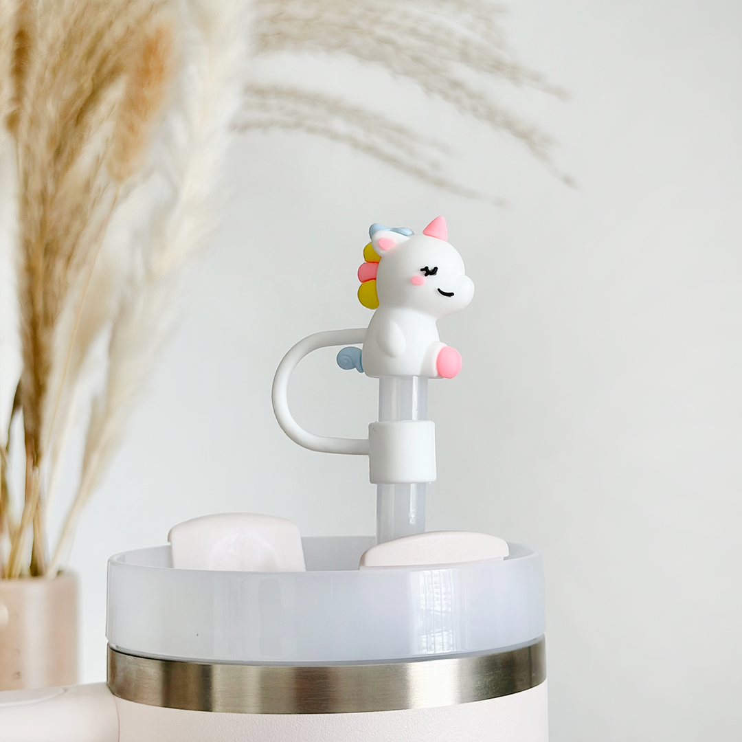 The Coconut Lane Unicorn Straw Cover, crafted from high-quality silicone, showcases a pink mane, pastel-colored horn, and small wings. Designed to fit on any white reusable water bottle with a stainless steel rim, this playful accessory is beautifully complemented by the elegant touch of decorative dried pampas grass stems in the background.