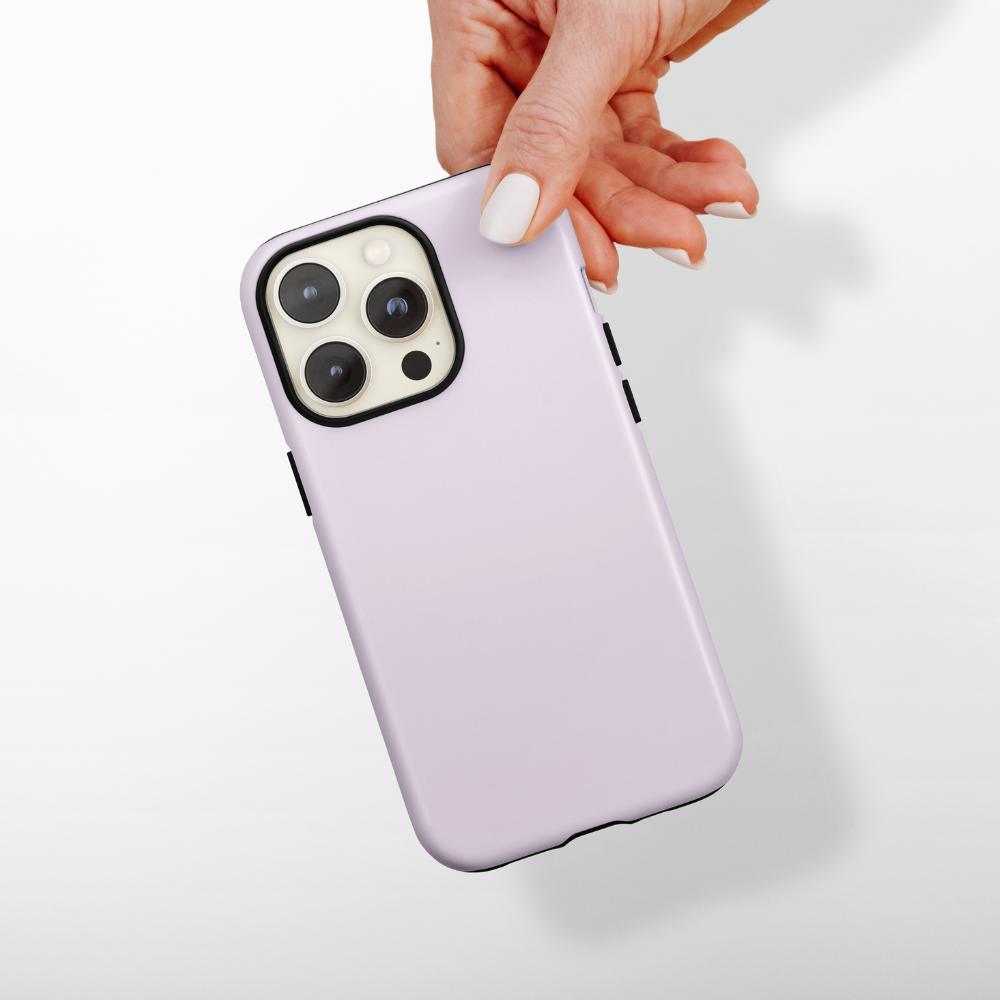 A hand with manicured nails is holding a smartphone with a light lilac "Tough Phone Case - NAKD Lilac" by Coconut Lane. The phone has a triple camera setup on the back. The background is plain and light-colored, casting a shadow of the hand and phone.
