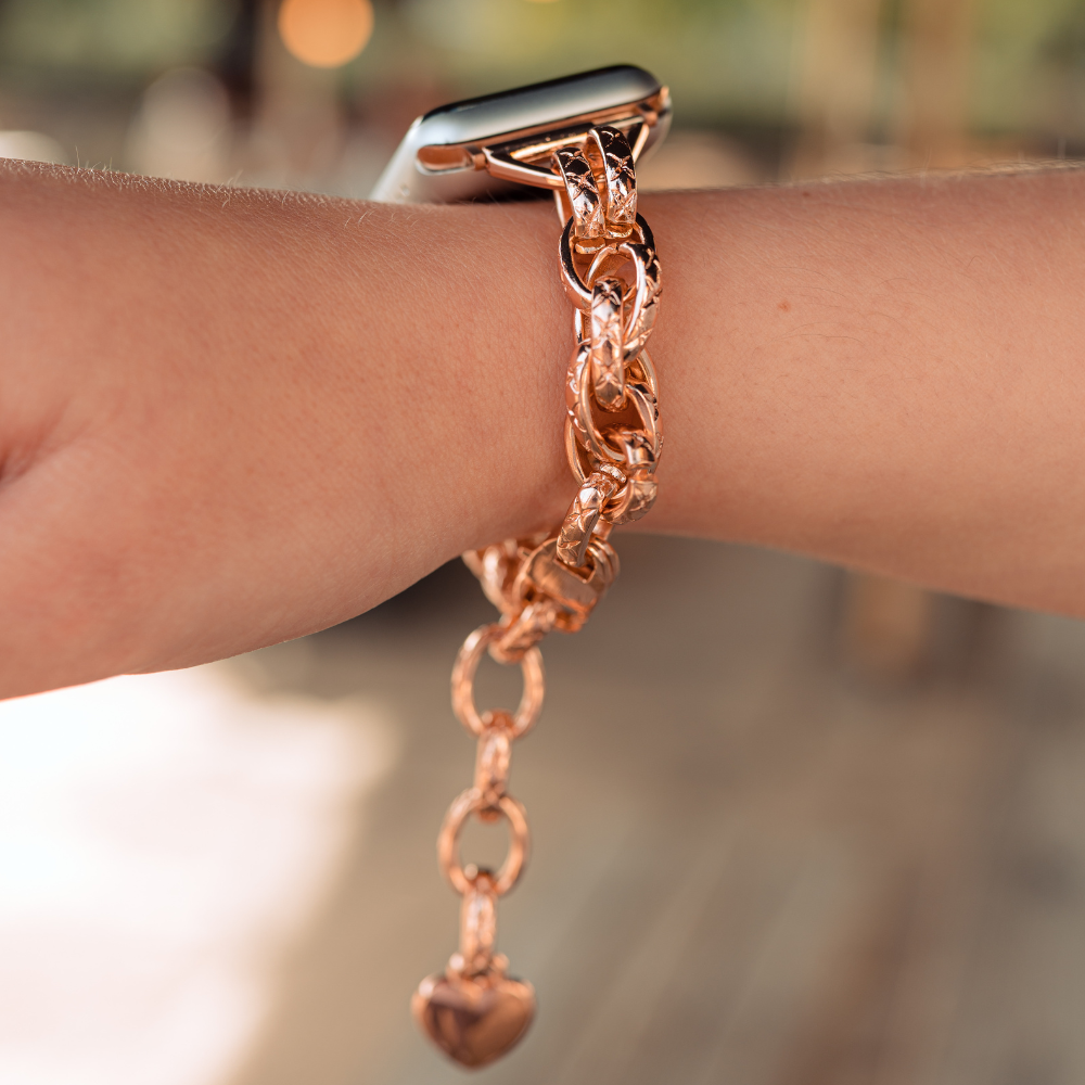 Close-up of a person's wrist showcasing the Chain Bracelet Apple Watch Strap - Rose Gold by Coconut Lane, featuring a heart-shaped charm. The person is stylishly pairing the rose gold strap with an Apple Watch, creating a cohesive and elegant look. The blurred background draws attention to these chic wrist accessories.