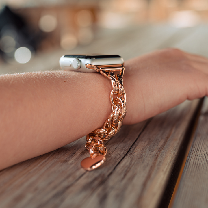 A wrist adorned with the Coconut Lane Chain Bracelet Apple Watch Strap in Rose Gold, featuring a stainless steel adjustable band with a chain-link design, resting on a wooden surface. The background is slightly blurred, drawing attention to the stylish accessory.