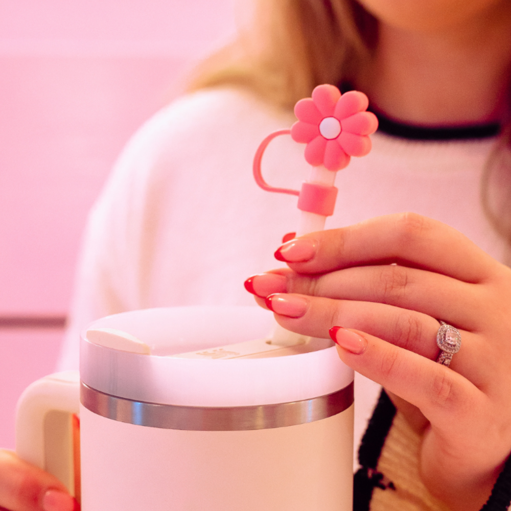 A person wearing a white sweater with black trim adjusts a Coconut Lane Pink & White Daisy Straw Cover on the lid of a white insulated travel mug. The individual has long, painted nails and is wearing a diamond ring on their ring finger. The background is pink.