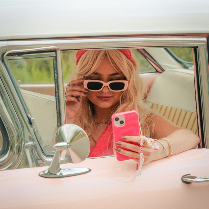 A person with long blond hair wearing a pink headband and white-framed sunglasses is sitting in a vintage car with a pink interior. They are taking a selfie with a Melting Heart Phone Case - Bright Pink from Coconut Lane, adorned with beaded charms, and holding the sunglasses.