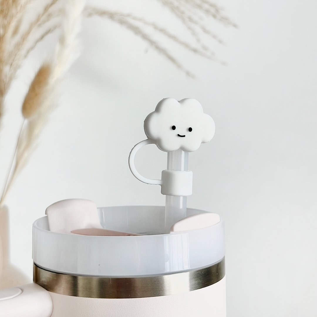 A close-up of a light pink tumbler with a silver accent. The tumbler features a white straw topped with Coconut Lane's adorable White Cloud Straw Cover accessory. In the background, there are blurred beige pampas grass against a light, neutral wall.