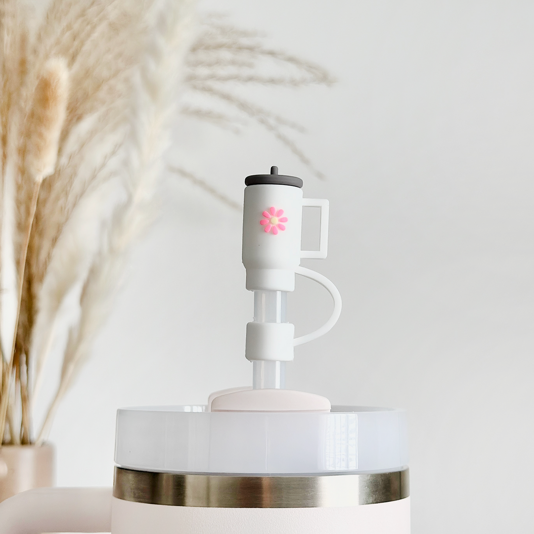 A Mini White Tumbler Straw Cover from Coconut Lane, adorned with a floral design on its spout, releases mist into the air. The device, used in conjunction with a silicone straw cover for added hygiene, is set on a smooth, pale surface. Dried pampas grass in a vase in the background enhances the soft, minimalist aesthetic.