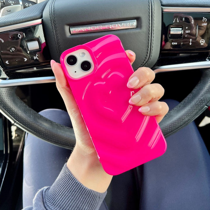 A hand with neatly manicured nails holds a pink phone encased in Coconut Lane's extra protective, bright pink Melting Heart Phone Case that features a 3D melting heart effect. The person is seated in a Range Rover, with the steering wheel displaying the Range Rover logo visible in the background.