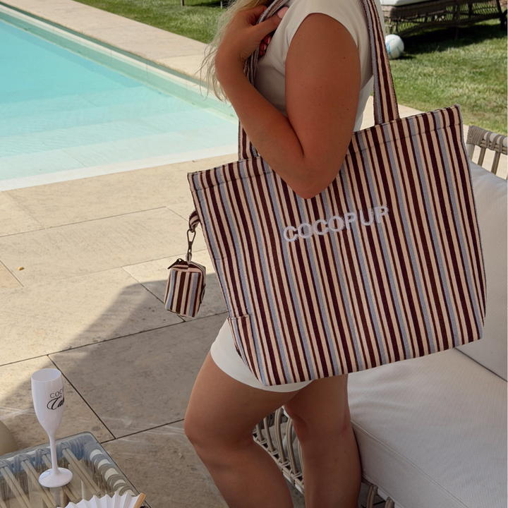 Woman poolside with a brown and blue striped tote bag, wearing white; drink set nearby.