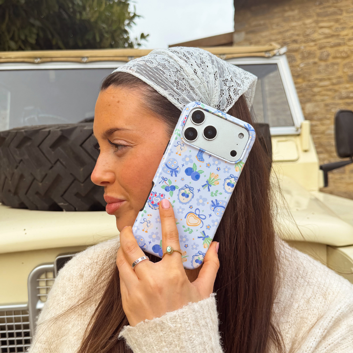 Girl wearing cream cardigan holding her blueberry picnic phone case next to cream car