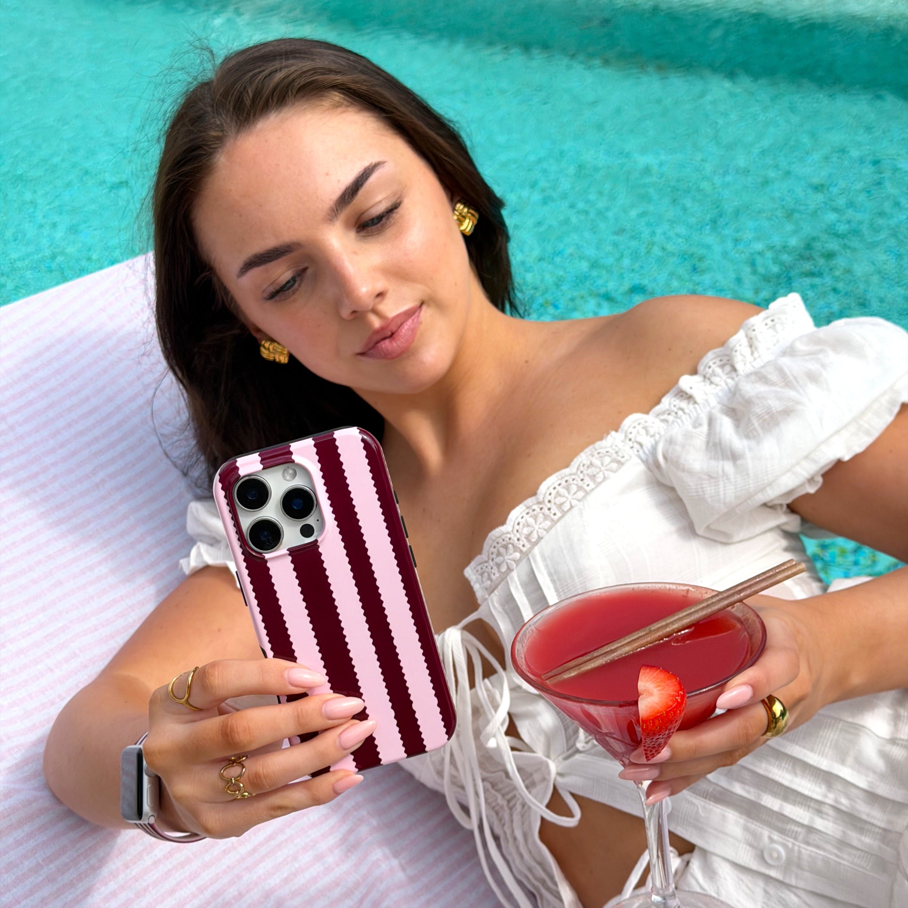 Girl holding a blushberry striped phone case, with strawberry daiquiri by the pool 
