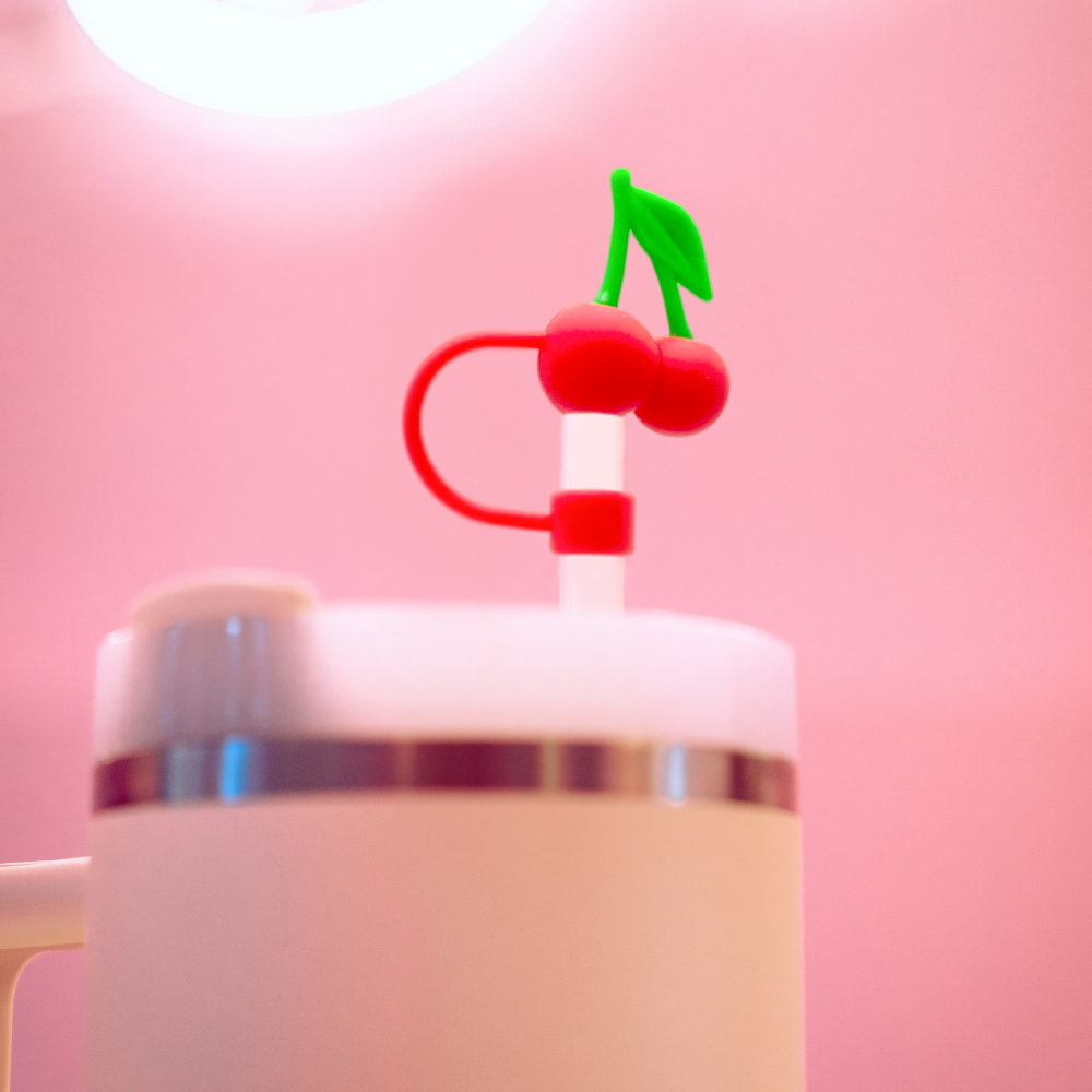 The "Cherries Straw Cover" by Coconut Lane is a white cup with a lid that features a straw topped with a whimsical cherry decoration and includes a high-quality silicone straw cover. Set against a blurred, soft pink background, the image evokes a playful and vibrant feel while ensuring hygiene.