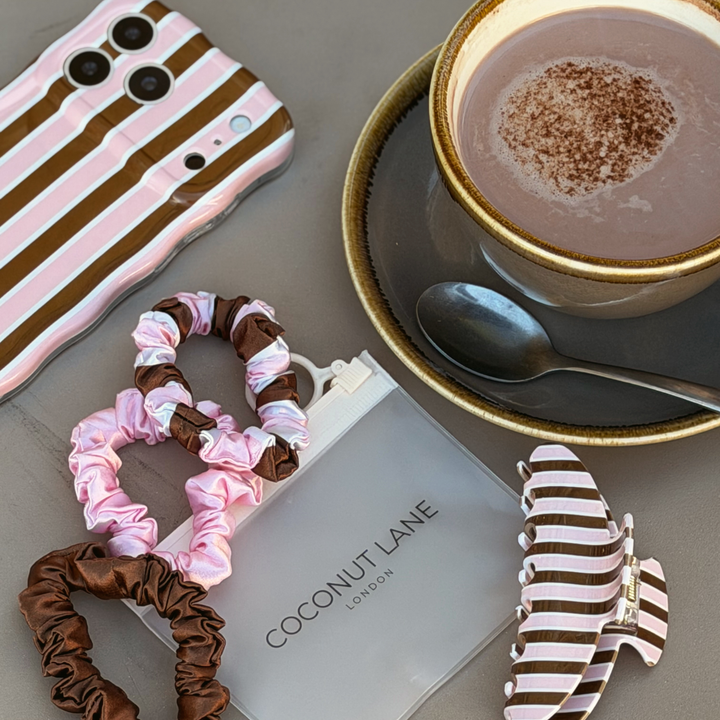 flat lay image of 3 satin scrunchies in a chocolate brown, pink and brown, pink and white striped design. Placed alongside the matching wavy phone case and claw clip.