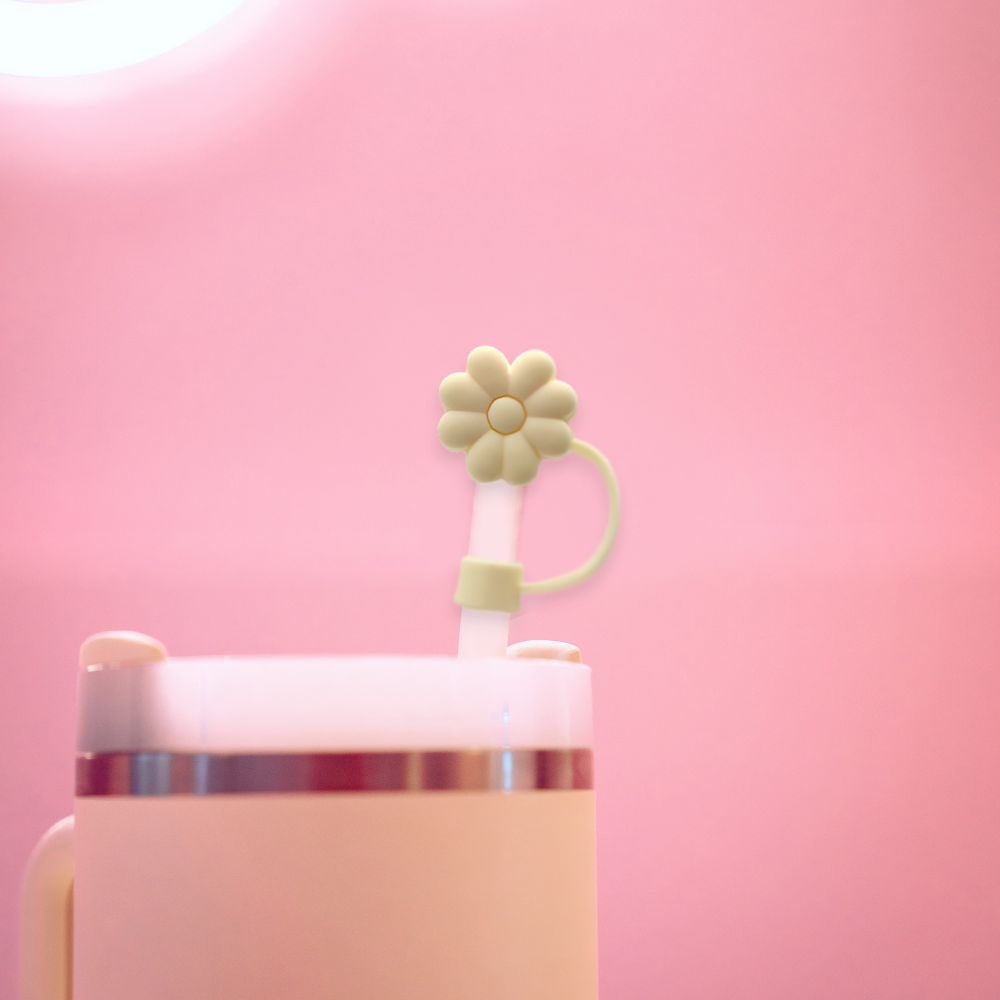 A white beverage tumbler with a soft pink lid and a straw topped with Coconut Lane's Cream Flower Straw Cover against a pastel pink background. The flower accent adds a cute and decorative touch to the beverage container.