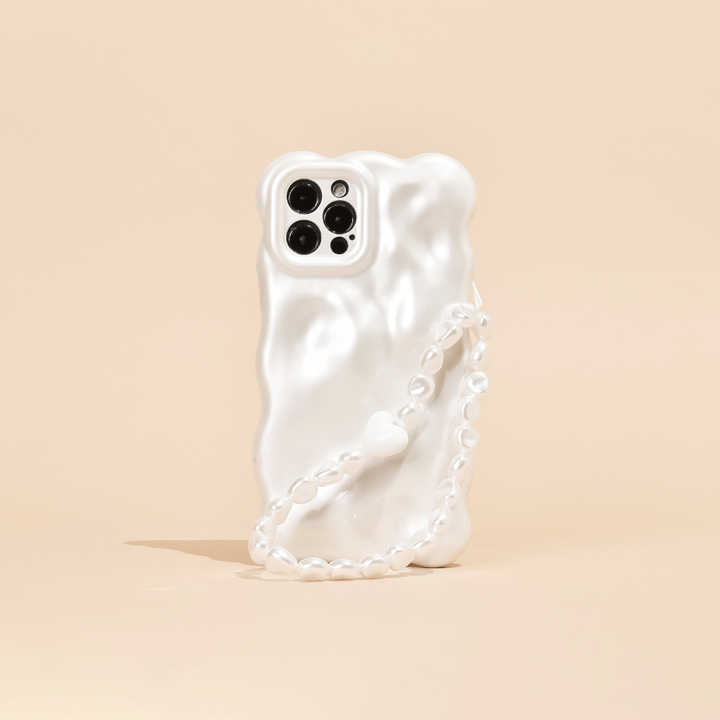 A white, wavy-textured phone case is shown standing on a light beige surface, featuring Coconut Lane's Beaded Phone Strap - Chunky Pearls. The phone's camera section with multiple lenses is visible, contributing to a sleek and modern aesthetic.