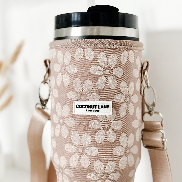 A beige fabric Tumbler Carry Case, named "Mocha Flower," from Coconut Lane, displays a delicate white floral pattern. It includes a metal lid and a shoulder strap. The softly blurred background highlights the elegance of the case.