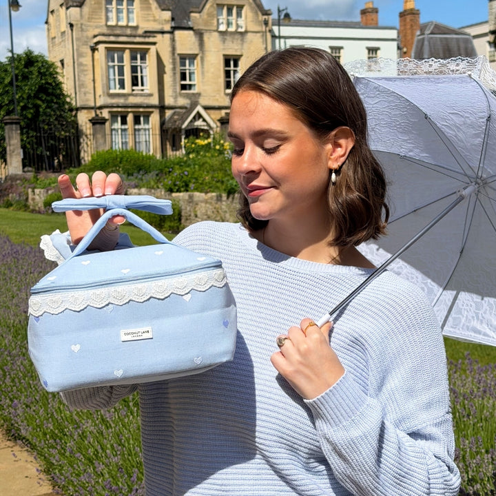 Woman holding blue vanity case with lace details and an umbrella in a garden setting.