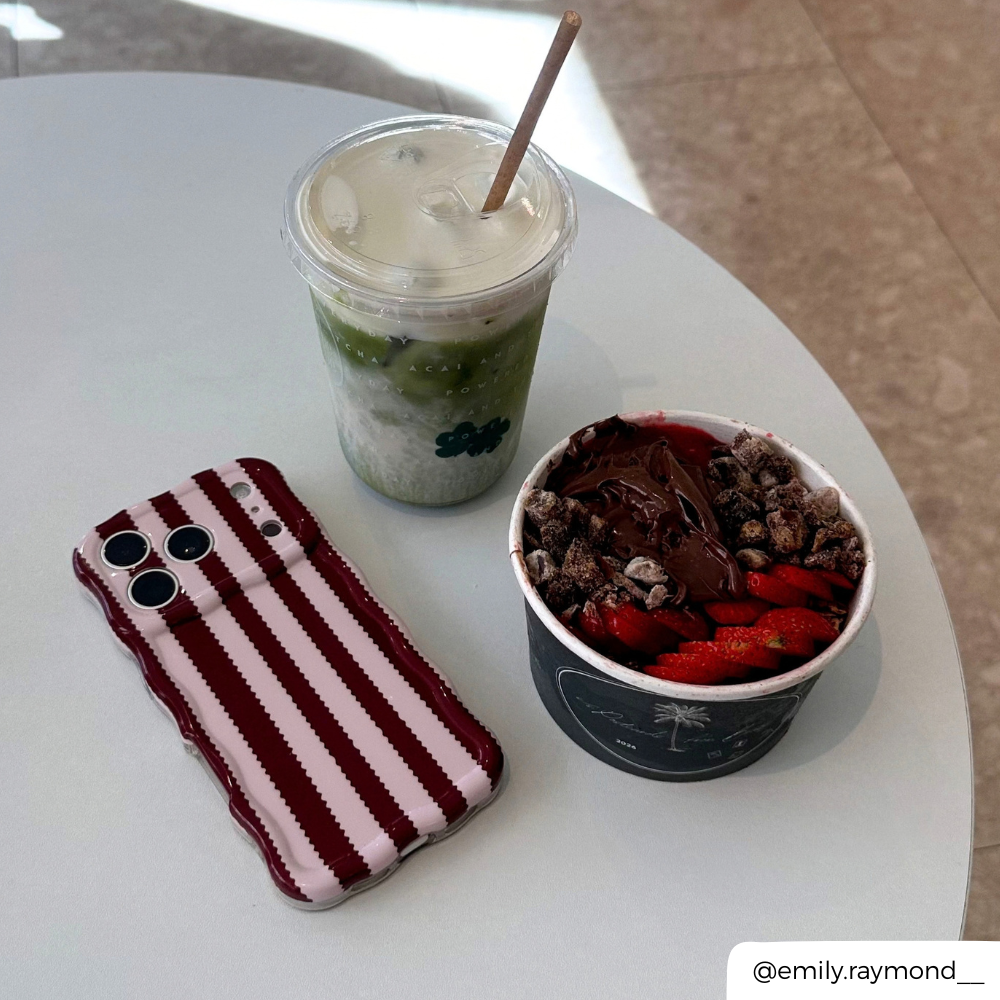 Iphone on table next to matcha and acai bowl. The phone is in a burgundy and light pink striped wavy phone case.