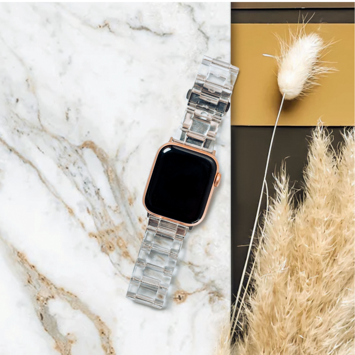 A smartwatch featuring the Luxe Transparent Apple Watch Strap by Coconut Lane and a black screen is placed on a light marble surface. To the right, there is a bundle of beige, dried pampas grass set against a dark background accented with mustard yellow tones.