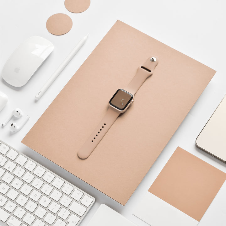 A beige smartwatch with a square face sits on a beige notebook, surrounded by an array of white office essentials such as a keyboard, mouse, pen, earphones, and laptop. Two round beige coasters are also visible. The NAKD Apple Watch Strap - Cappuccino by Coconut Lane adds an elegant touch to the setup.