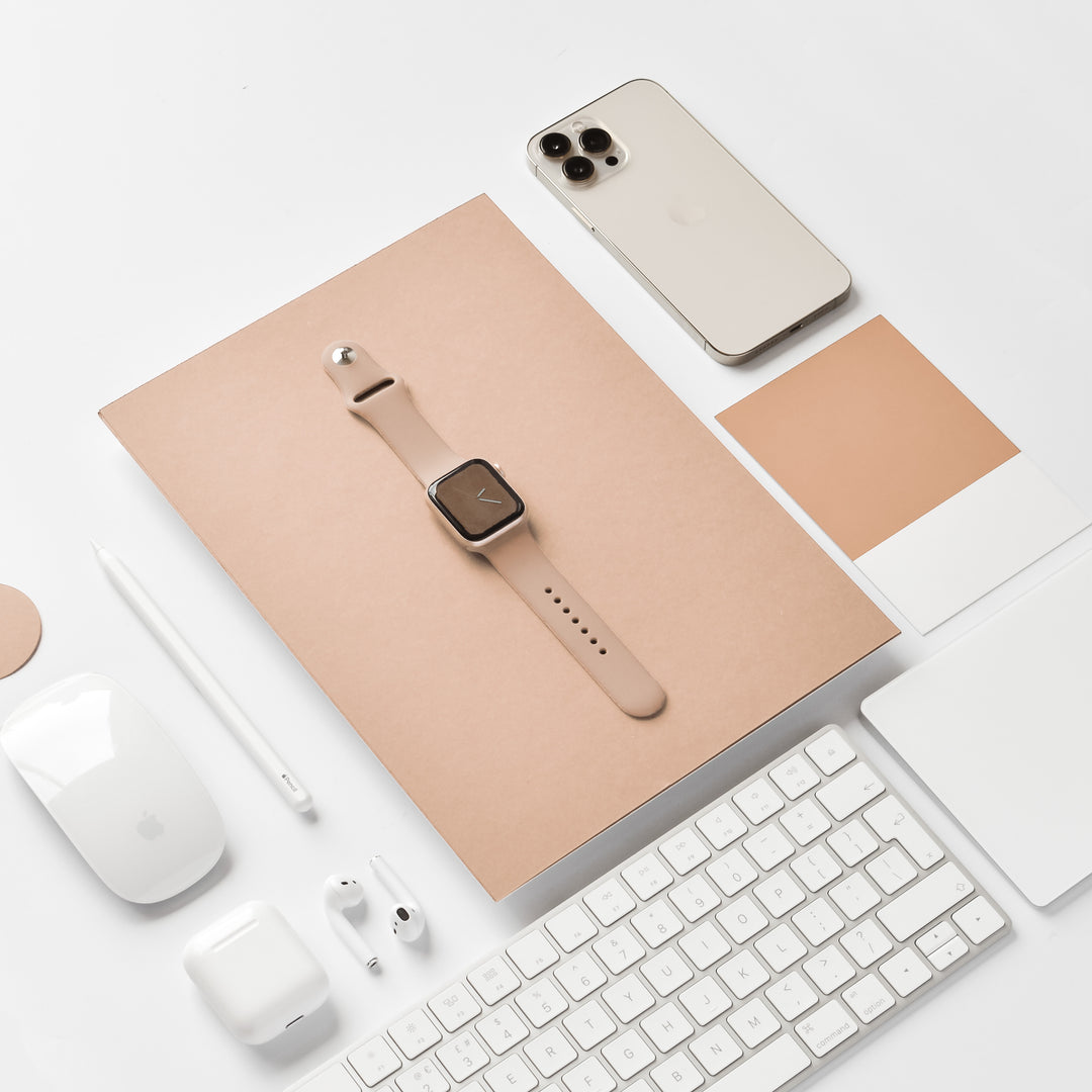 A neatly arranged workspace featuring a smartwatch with a NAKD Apple Watch Strap - Cappuccino by Coconut Lane, a beige smartphone, white wireless earbuds, a white stylus, a white computer mouse, a white keyboard, a white notebook from the NAKD Collection, and color swatches in beige tones on a beige notepad.