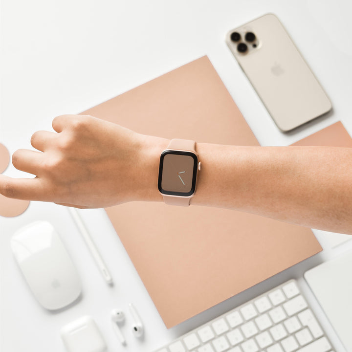 A person is wearing the Coconut Lane NAKD Apple Watch Strap in Cappuccino on their wrist. The background features an Apple iPhone, a wireless mouse, a stylus, earphones, a keyboard, and a notebook, all in beige and white tones. This minimalist tech setup is enhanced by the stylish NAKD Collection silicone Apple Watch Bands from Coconut Lane.