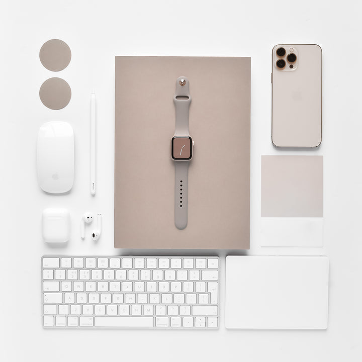 A neatly arranged collection of beige and white tech accessories, part of the NAKD Collection by Coconut Lane, includes an Apple Watch with a NAKD Apple Watch Strap in Mocha, an iPhone, a MacBook, a Magic Mouse, a keyboard, a stylus, AirPods, and a touchpad. All items are placed on a white background in a symmetric and aesthetically pleasing manner.