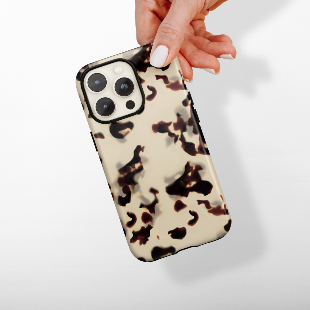 A hand with white painted nails holds the Coconut Lane Tough Phone Case in Ivory Tort. The beige and black tortoiseshell-patterned case provides 360-degree protection for the smartphone, with its shock absorbent design ensuring safety against a white background, prominently displaying the camera lenses.