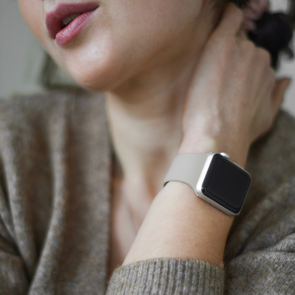 A close-up shows a person with fair skin and light pink lipstick, donned in a grey cardigan. They have a silver smart watch on their left wrist, featuring the NAKD Apple Watch Strap - Caramel Latte from Coconut Lane, positioned near their neck. The background is blurred.