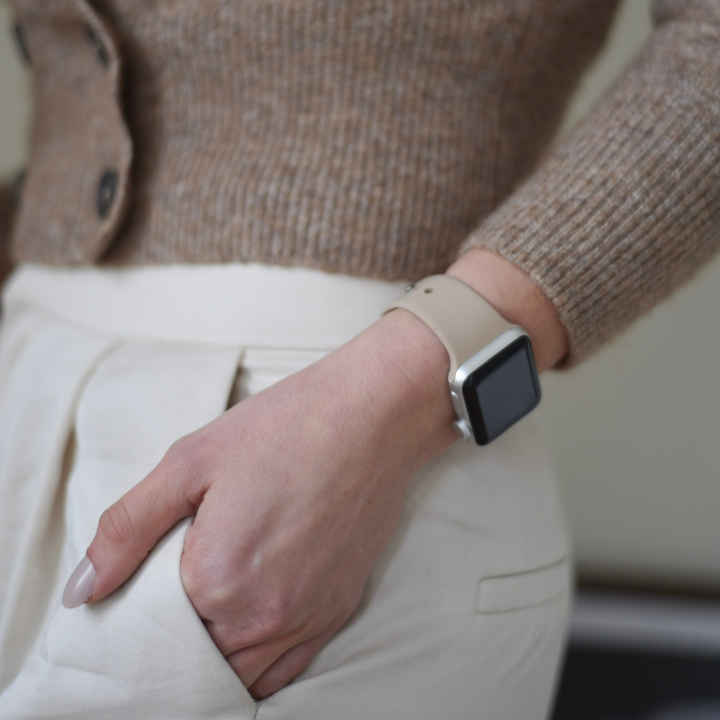 A close-up shot features a person's left hand, adorned with neatly manicured nails, as it rests in the pocket of cream-colored trousers. The individual is donning a beige sweater and showcasing the Coconut Lane NAKD Apple Watch Strap in Caramel Latte on their wrist. The background is blurred, directing attention to the hand and the elegant watch strap.