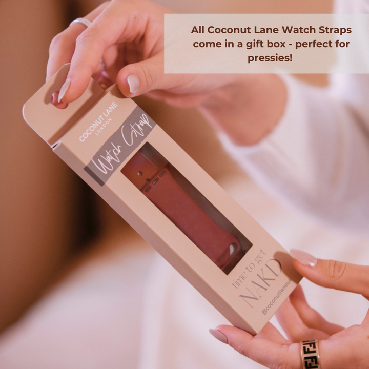 A person is holding a box containing a NAKD Apple Watch Strap in Red Wine, branded "Coconut Lane." The box features a window displaying the strap and reads "time to get NAKD." A text overlay states, "All Coconut Lane silicone straps come in a gift box - perfect for pressies!