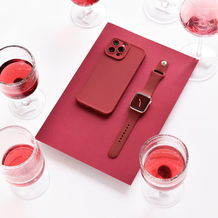 A red smartphone and a matching Apple Watch with a NAKD Apple Watch Strap in Red Wine by Coconut Lane are placed on a red rectangular surface. Surrounding them are six glasses filled with red liquid, giving a sophisticated and coordinated aesthetic.