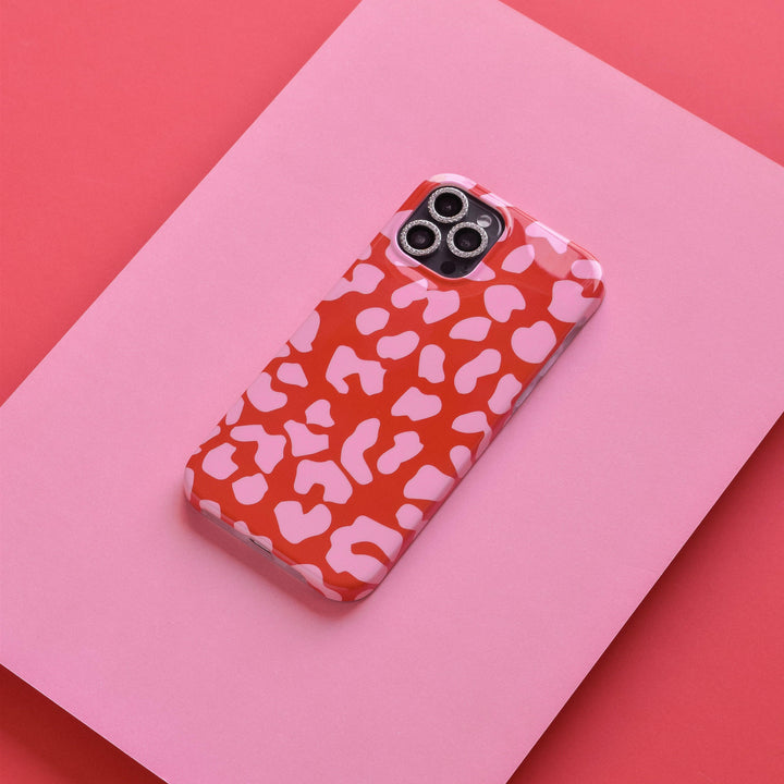 A smartphone with a red and pink animal print case, featuring Coconut Lane's Diamante Protective Camera Lens Covers and scratch-resistant tempered glass, rests on a larger pink paper backdrop, which in turn is placed on an angled red surface. The combination of colors and patterns creates a visually striking contrast.