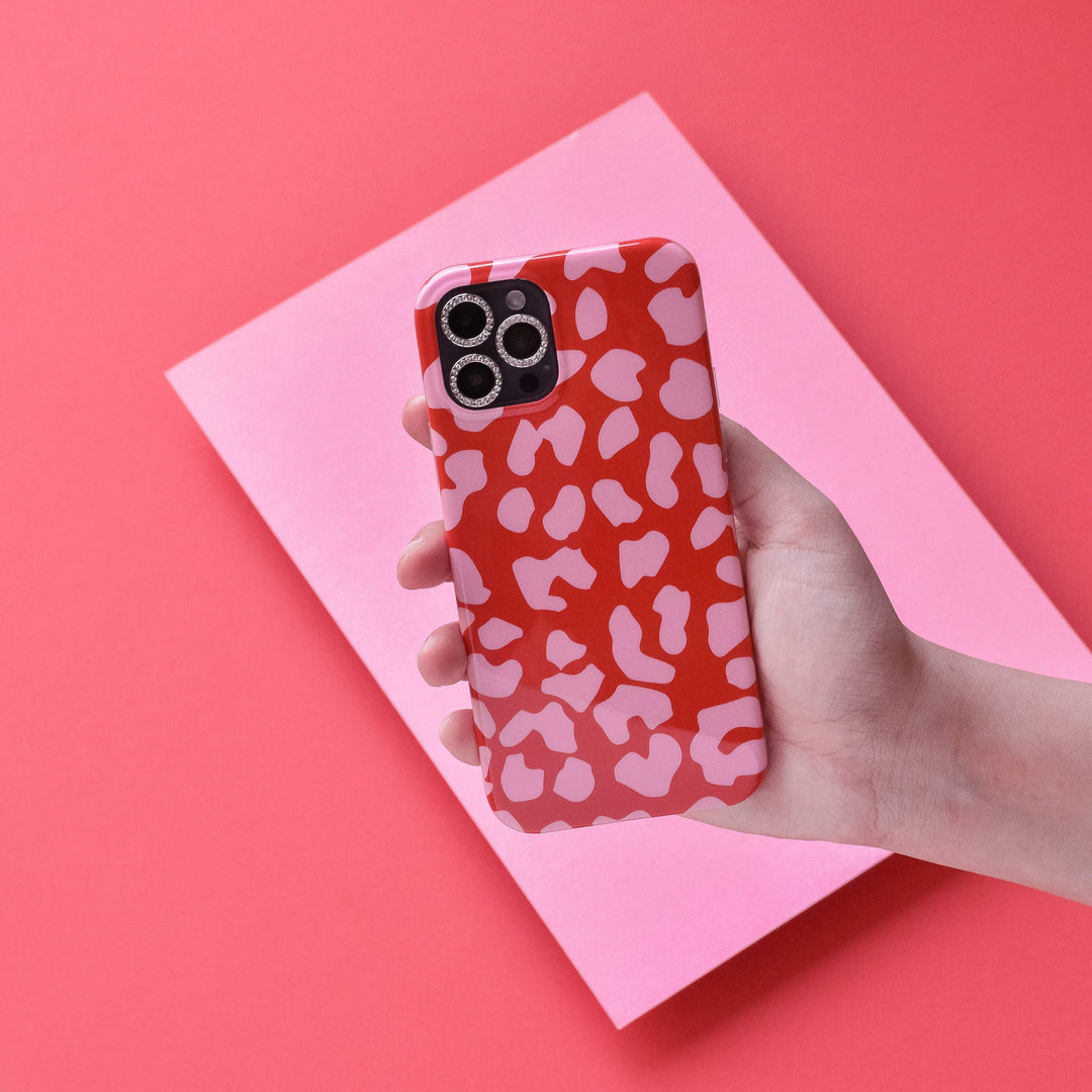 A hand holding an iPhone with a red and pink patterned case against a pink and red background. The phone's camera lenses are visible, protected by Coconut Lane's tempered glass "Protective Camera Lens Covers - Diamante," while the scratch-resistant case features abstract heart-like shapes.