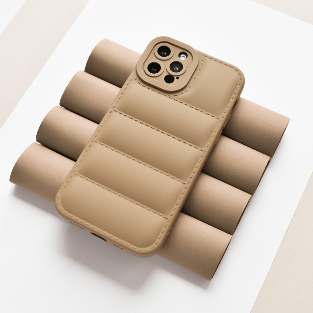 The Quilted Phone Case - Brown by Coconut Lane is displayed lying face down on a set of brown cylindrical objects, showcasing its full camera protection. Made from soft, flexible TPU, the background features a neutral palette with white, beige, and light gray tones.