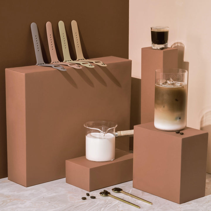 A minimalistic setup features brown blocks arranged at different heights, showcasing beverage items including a glass with iced coffee and another with milk. A small shot of espresso and several coffee spoons are also present. Five NAKD Apple Watch Straps in Mocha from Coconut Lane hang on a block.