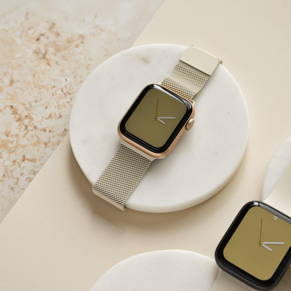 A Magnetic Chain Apple Watch Strap - Almond White by Coconut Lane, featuring a gold finish with a beige mesh band and a stainless steel adjustable clasp, is elegantly displayed on a round white marble platform. Another similar smartwatch can be seen partially in the background on a circular white surface. The composition, embodying metallic minimalism, rests on a textured beige surface, creating an elegant and sophisticated setup.