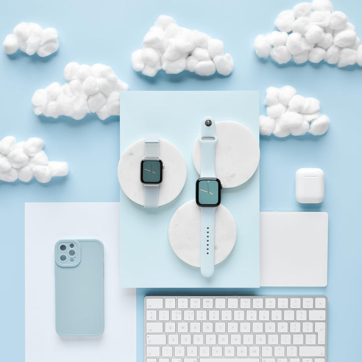 A flat lay of smartwatch and tech accessories includes two smartwatches with pastel bands on marble coasters, a Coconut Lane NAKD Phone Case in baby blue featuring soft flexible TPU for full camera protection, a white keyboard, a computer mouse, and an AirPods case. The background is light blue with fluffy white cotton clouds.