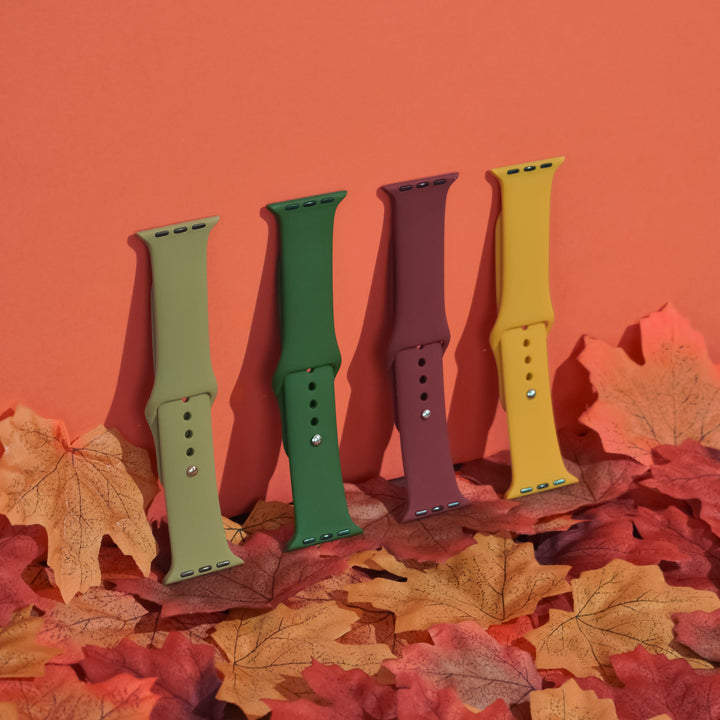 Four Coconut Lane's NAKD Apple Watch Straps in green, dark green, burgundy (Red Wine), and mustard yellow are positioned vertically in a row against a red background. They feature silicone straps and are surrounded by scattered autumn leaves in shades of orange and yellow.