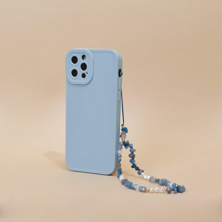 A light blue smartphone case with a camera cutout is displayed standing vertically against a beige background. Attached to the case is Coconut Lane's "Beaded Phone Strap - Ocean Eyes," made of blue beads and small pearls, adding an elegant touch to your phone accessories.