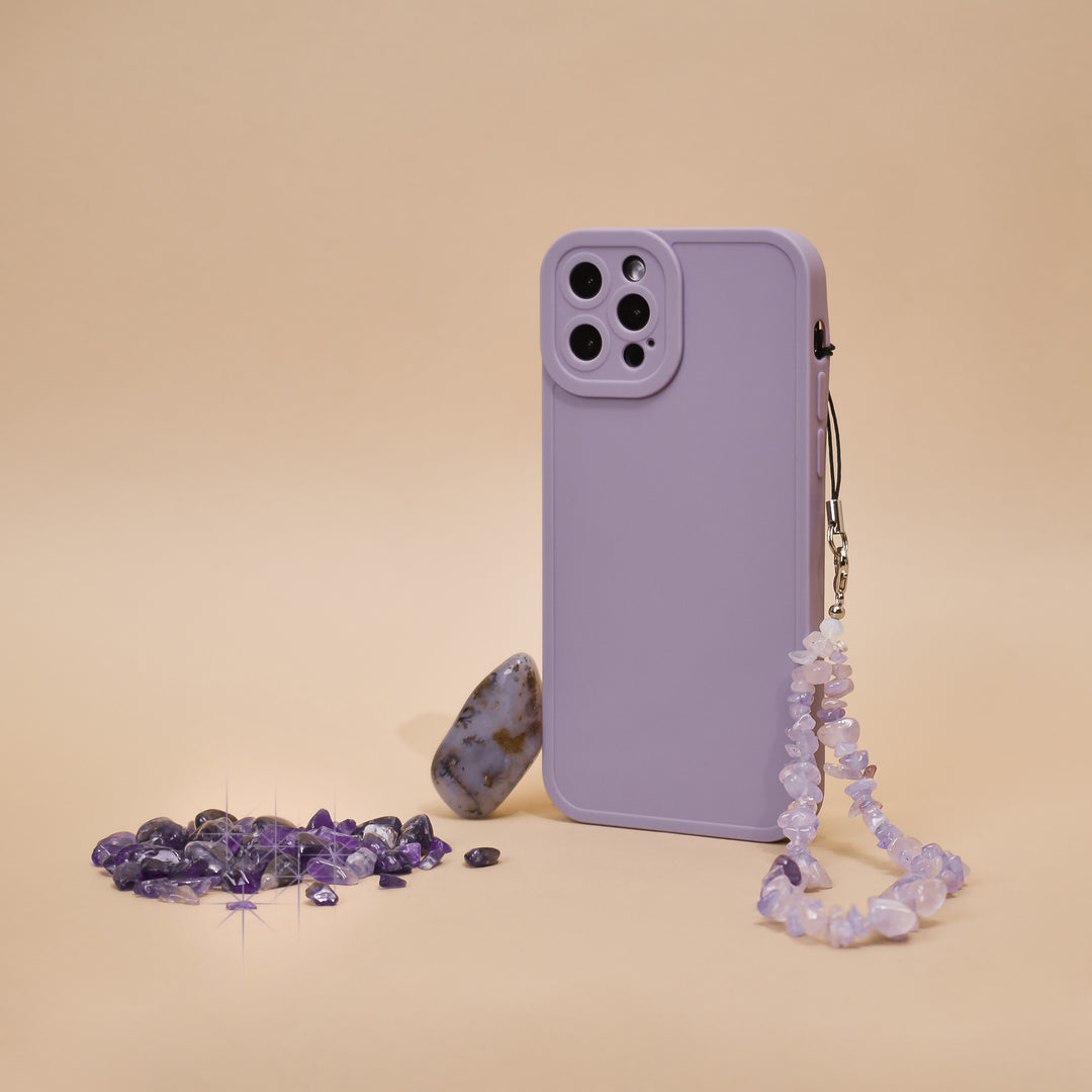 A phone case in a vibrant lilac shade with Coconut Lane's Crystal Phone Strap adorned with small beads stands upright against a beige background. Scattered around the stylish accessory are amethyst crystals and one larger, smooth stone with earthy tones.